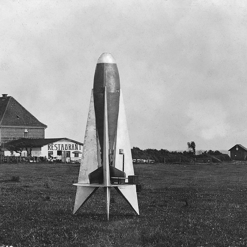 Winkler-Rakete HW-2 auf der Greifswalder Oie, ca. 1931. BN_14512. Rakete steht senkrecht auf einer Wiese, im Hintergrund Gebäude und das Meer.